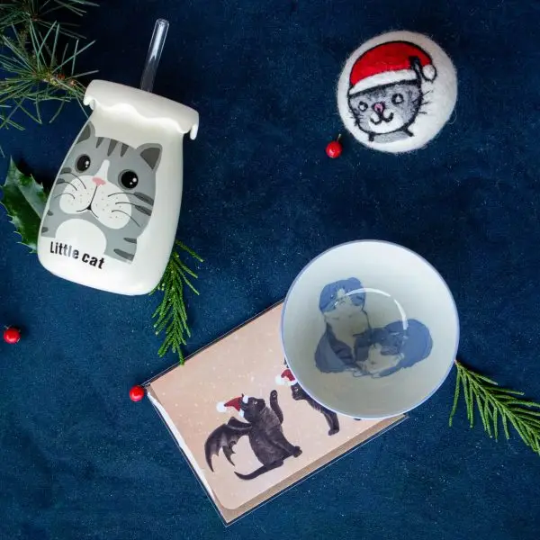 Gifts for cat lovers. Drinking bottle with straw and cat illustration that says little cat. Wool Dryer ball with cat wearing a santa hat felted onto it. Greeting card with two black cats with bat wings and santa hats illustration. Small ceramic bowl with blue cat illustration.