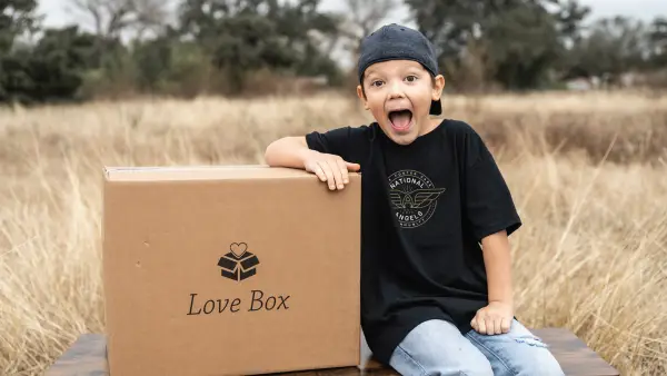 You boy wearing a Salem Angels shirt poses with a large cardboard box that says Love Box