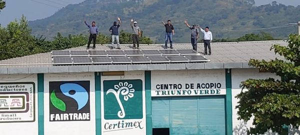 A group of men on a roof with solar panels, with a sign on the wall reading "Centro de Acopio, Triunfo Verde"