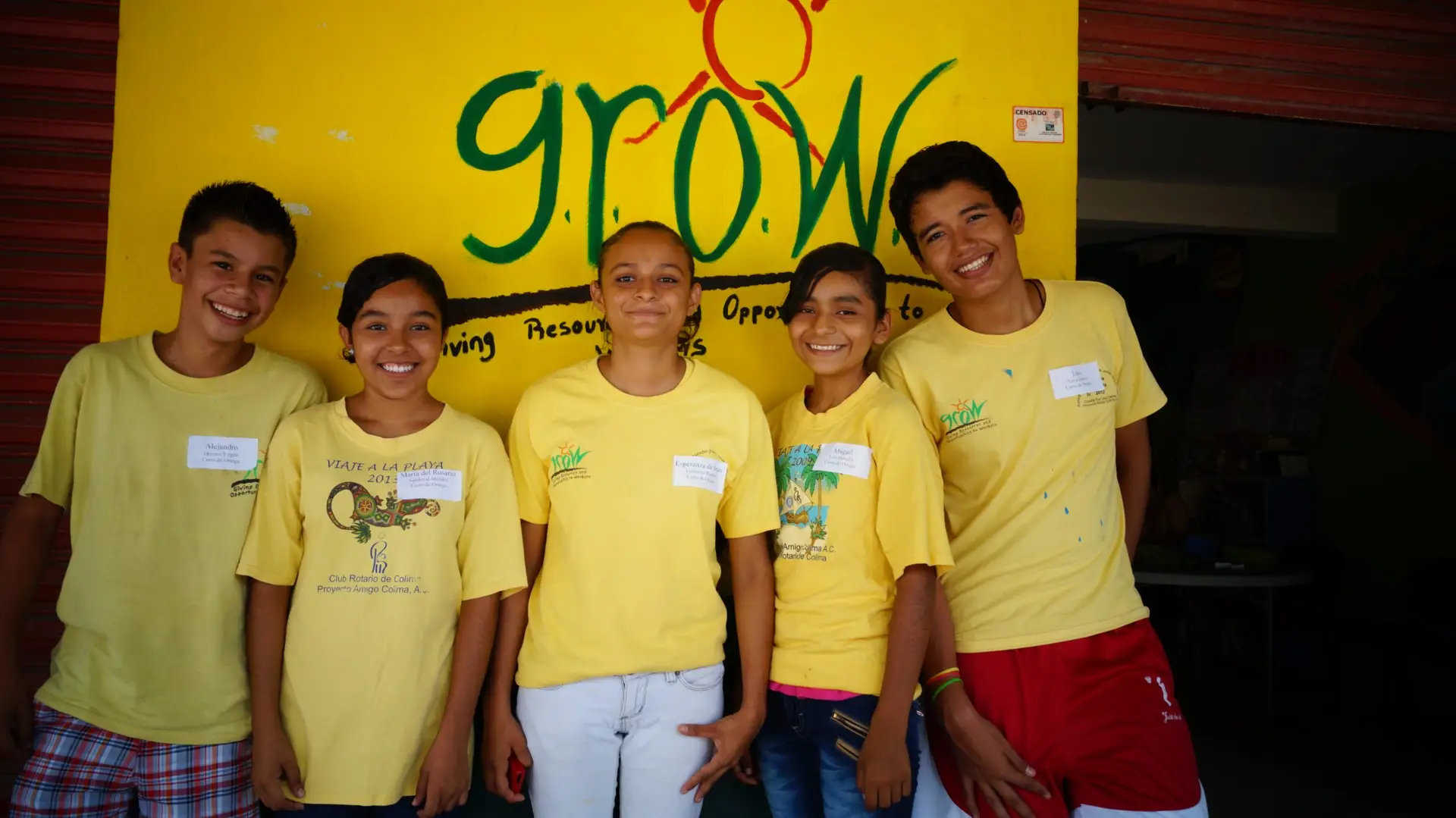Five smiling students wearing yellow GROW program shirts stand in front of a bright yellow GROW sign in Colima, Mexico, representing educational opportunities funded through fair trade banana purchases.