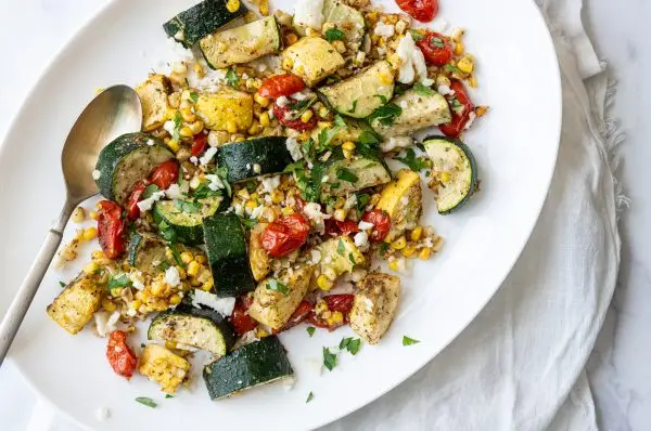 Roasted zucchini, yellow squash, sweet corn, and cherry tomatoes on a white platter, topped with fresh parsley and crumbled feta cheese.