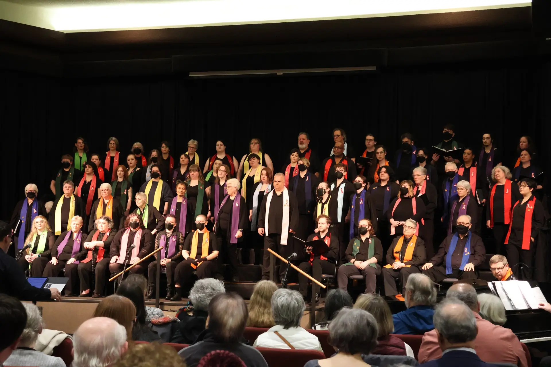 Members of Confluence: Willamette Valley LGBT Chorus perform on stage before a seated audience. Singers of varying ages stand on risers wearing black attire with colorful rainbow scarves, celebrating community, diversity, and LGBTQ+ pride through music.