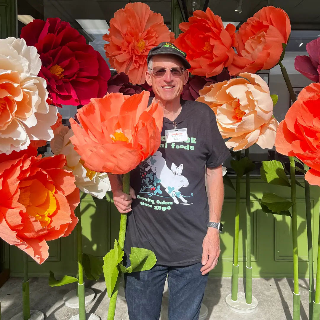 LifeSource founder Alex smiling among oversized orange, red, and cream paper flowers outside the store during Food Fair.