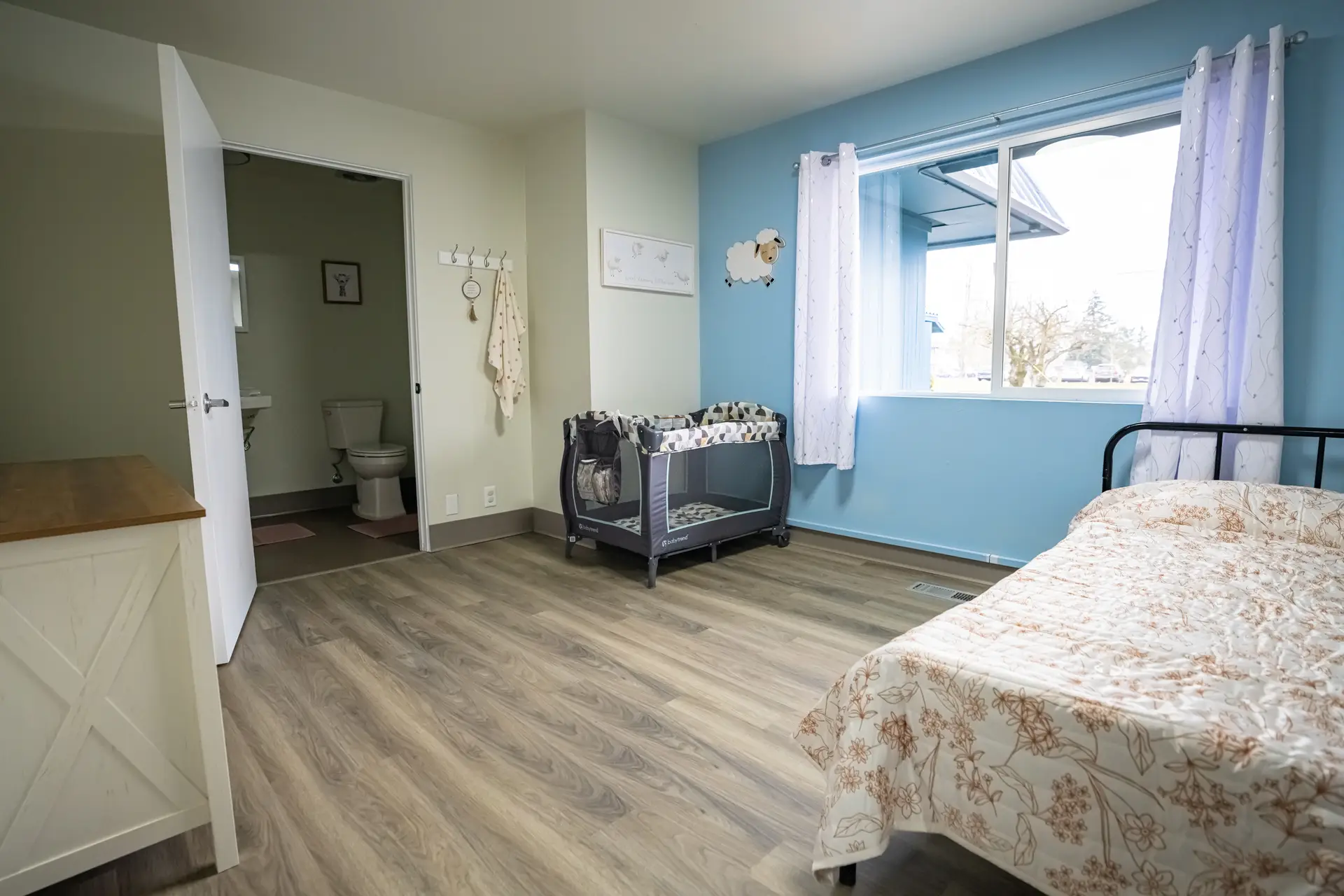 A furnished family shelter room with a bed, crib, private bathroom, and large window.