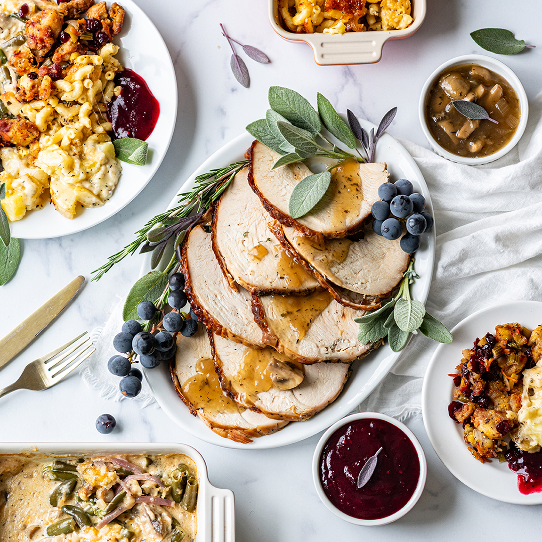 Sliced roasted turkey breast with mushroom gravy, surrounded by scratch-made holiday sides including stuffing, mac and cheese, cranberry sauce, potatoes, and green bean casserole.