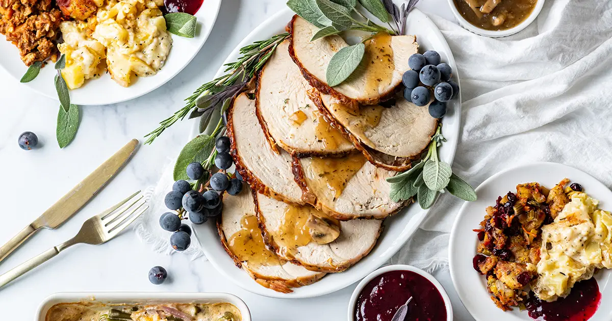 Sliced roasted turkey breast with mushroom gravy, surrounded by scratch-made holiday sides including stuffing, mac and cheese, cranberry sauce, potatoes, and green bean casserole.